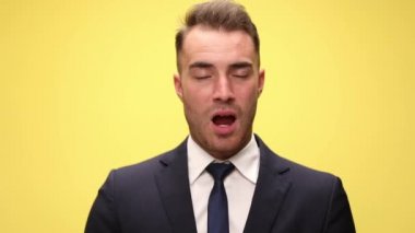 Uykulu, takım elbiseli genç iş adamı ağzını açıyor ve esniyor. Ağzını eliyle kapatıyor ve sarı arka planda gülümsüyor.