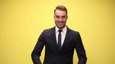 Mutlu, zarif takım elbiseli iş adamı gülümsüyor, kollarını açıyor, sarı arka planda hoş karşılıyor ve gülümsüyor.