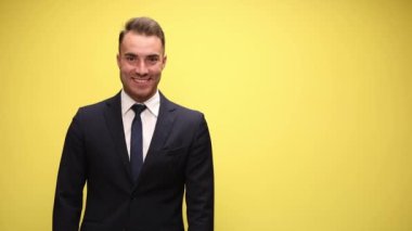 Takım elbiseli mutlu genç işadamı gülümsüyor, bakıyor ve sarı arka planda sunuyor.