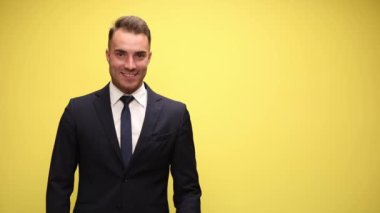 Takım elbiseli mutlu genç manken yan yana bakıyor ve sunuyor, parmaklarını havaya kaldırıyor ve zaferi kutluyor, sarı arka planda gülümsüyor.