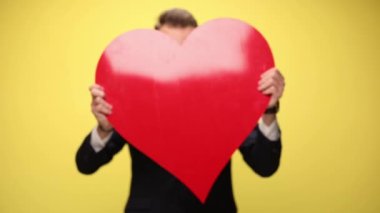 Büyük kırmızı kalbin arkasına saklanan şık takım elbiseli mutlu iş adamı, sarı arka planda kalbini gösteriyor ve sunuyor.