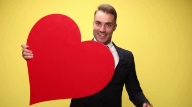 Şık takım elbiseli genç iş adamı büyük kırmızı bir kalp tutuyor, gülümsüyor ve bana jest diyor, hareket ediyor, dans ediyor ve sarı arka planda el kol hareketleri yapıyor.