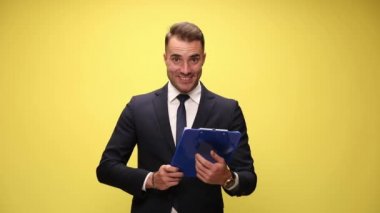 Takım elbiseli genç işadamı elinde not defteri, gülümsüyor ve sarı arka planda baş parmak hareketi yapıyor.