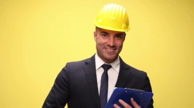 Güvenlik kaskı takarak gülümseyen zarif mühendis, panodan okuyor ve sunuyor, el ele tutuşuyor ve sarı arka planda takım elbise ayarlıyor.