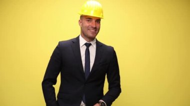 Gülümseyen mühendis takım elbise ve güvenlik kaskı takmış yukarı bakıyor, gülümsüyor ve sarı arka planda parmaklarını işaret ediyor.