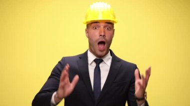 Güvenlik kaskı takan mutlu, zarif mühendis, yukarı bakıyor, haberleri duyuyor, elleriyle yüzünü örtüyor, ağzını açıyor ve elini havada tutuyor sarı arka planda zaferi alkışlıyor.