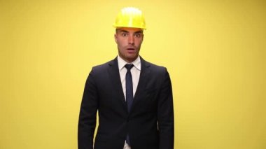 Şaşırmış mühendis, güvenlik kaskı takıyor, yukarı bakıyor, haberleri dinliyor, ağzını açıyor ve yüzünü avuçluyor, kaskını fırlatıyor ve sarı arka planda duruyor.
