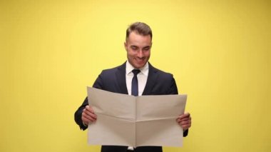 şaşırmış genç işadamı gülümsüyor, gazete okuyor ve sarı arka planda ağzını açıyor.