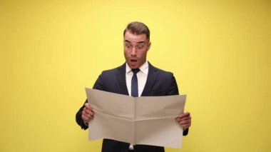 Takım elbiseli şık bir işadamı gazete okuyor, şok edici bir şekilde ağzını açıyor ve sarı arka planda gazeteyle yüzünü örtüyor.