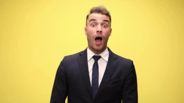 Takım elbiseli genç iş adamı haberleri dinliyor, ağzını açıyor, parmaklarını çapraz yapıyor ve sarı arka planda şans işareti yapıyor.