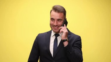 Takım elbiseli şık bir işadamı telefonda konuşuyor, gülümsüyor ve sarı arka planda güzel bir jest yapıyor.
