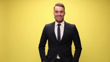 Mutlu, zarif takım elbiseli iş adamı gülümsüyor, el ele tutuşuyor, yüzüne ve çenesine dokunuyor ve sarı arka planda düşünüyor.