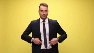 Mutlu genç zarif iş adamı ceketini ilikliyor ve kravatı ayarlıyor, gülümsüyor ve parmaklarını işaret ediyor, sarı arka planda duruyor.