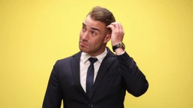 Takım elbiseli cahil genç işadamı başını kaşıyıp düşünüyor, bir fikri var ve onu işaret ediyor, gülümsüyor ve sarı arka planda duruyor.