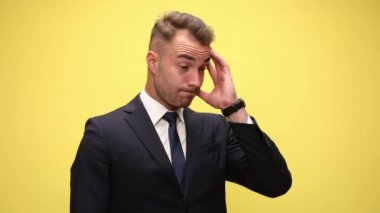 Şaşkın, şık takım elbiseli bir iş adamı etrafa bakıp kafa karıştırıyor ve düşünüyor, bir fikri var ve parmağıyla işaret ediyor, gülümsüyor ve sarı arka planda duruyor.