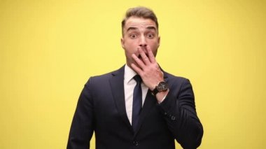 Şok olmuş şık takım elbiseli iş adamı haberleri duyuyor ve elleri ağızla yüzünü kapatıyor, gülümsüyor ve sarı arka planda duruyor.