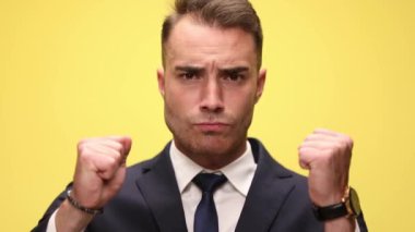 Şans dileyen takım elbiseli genç, zarif bir işadamı, yumruklarını kaldırıyor, el ele tutuşuyor ve sarı arka planda bir zafer umuyor.