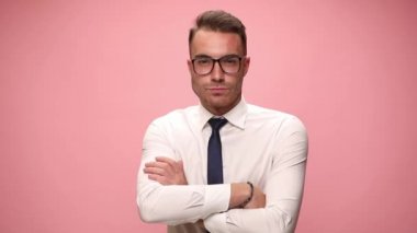 Gülümseyen beyaz gömlekli, gözlüklü, kollarını kavuşturup pembe arka planda duran gözlüklere bakan genç moda adamı.