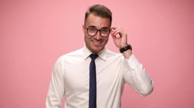 Beyaz gömlekli mutlu genç adam gülümsüyor, gözlüklere bakıyor ve pembe arka planda baş parmak hareketi yapıyor.