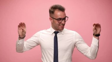 Beyaz gömlekli, gözlüklü, el ele tutuşup konuşan, her iki tarafa bakan, pembe arka planda gülen genç, zarif bir iş adamı.