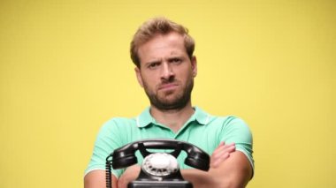 Genç adam kollarını kavuşturuyor, eski telefona bakıyor ve kaşlarını çatıyor, gülümsüyor ve sarı arka planda gülüyor.