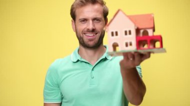 Kendine güveni tam, genç bir adamın evi kucaklayıp sunması, gülümsemesi, el sıkışması ve sarı arka plan anlaşmasını kabul etmesi.