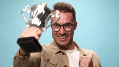 Gözlüklü ve ceketli çekici bir adam mavi arka plandaki başarılarını kutlamak için kameraya ödülünü sunuyor.