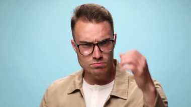 Genç kızgın adam kameraya yumruğunu gösteriyor, parmağını gösteriyor ve mavi arka planda tehditler savuruyor.