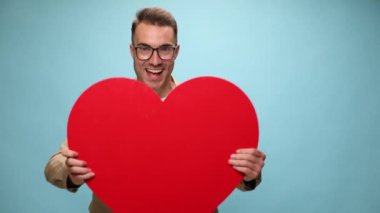 Yakışıklı, sıradan bir adam büyük kırmızı kalbini kameraya gösteriyor sonra sarılıyor ve mavi arka planda sırıtıyor.