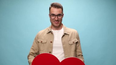 Çekici, sıradan bir adam yavaşça büyük kırmızı kalbini kaldırıyor ve mavi arka planda kameraya sunuyor.