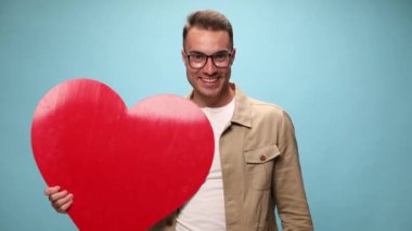 Genç, sıradan bir adam başparmağını kaldırıyor ve mavi arka planda kolunda büyük kırmızı bir kalp taşıyor.
