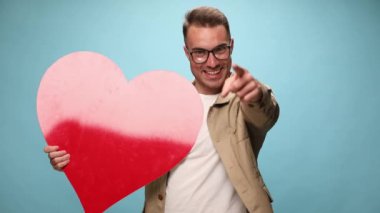 Büyük kırmızı bir kalp tutan seksi bir adam, kamerayı işaret ediyor, sonra onu işaret ediyor ve son olarak da kalbi, mavi arka planda başparmağıyla onaylıyor.