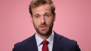 Güler yüzlü, lacivert takım elbiseli, kaşlarını kaldıran, yüzünü buruşturan, gülümseyen ve pembe arka planda sallanan zarif genç adam.