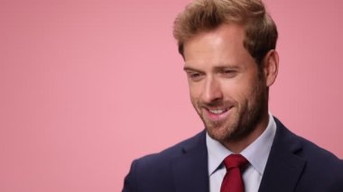 Lacivert takım elbiseli zarif bir adam kaşlarını kaldırıyor, flört ediyor, kravatını düzeltiyor ve pembe arka planda gülümsüyor.