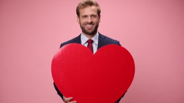 Mutlu, lacivert takım elbiseli, gülümseyen, kucaklayan ve pembe arka planda büyük kırmızı kalbi sunan zarif iş adamı.