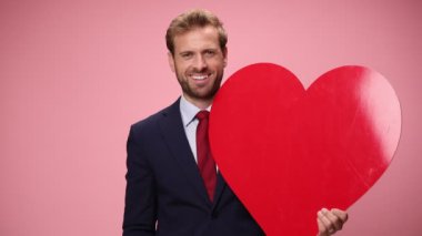 Lacivert takım elbiseli mutlu genç işadamı büyük kırmızı bir kalp tutuyor, parmaklarını gösterip pembe arka planda gülümsüyor.