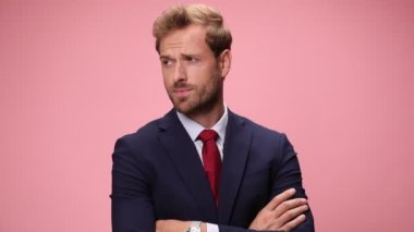 Lacivert takımlı seksi genç adam kollarını kavuşturuyor, çenesine dokunuyor, yan yana bakıyor, el ele tutuşuyor, takım elbisesini ayarlıyor ve gülümsüyor, pembe arka planda el ele tutuşuyor.