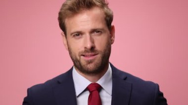 Lacivert takım elbiseli seksi genç işadamı gülümsüyor, kaşlarını kaldırıyor ve göz kırpıyor, pembe arka planda flört ediyor.