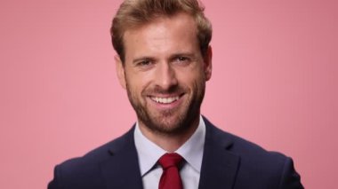 Deniz mavisi takımlı mutlu genç adam domuz kumbarası tutuyor, gülümsüyor ve pembe arka planı işaret ediyor.