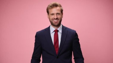 Lacivert takım elbiseli, kırmızı yüzlü ve pembe arka planda gülümseyen zarif genç iş adamı.