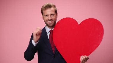 Lacivert takım elbiseli zarif genç işadamı büyük kırmızı bir kalp tutuyor, gülümsüyor, parmaklarını işaret ediyor ve pembe arka planda baş parmak hareketi yapıyor.