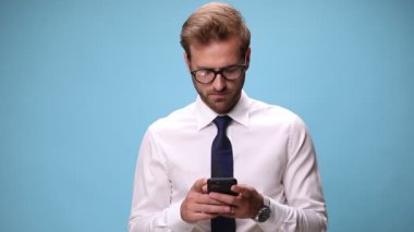 Genç işadamı telefonuyla mesajlaşıyor, gülüyor ve mavi arka planda onay veriyor.