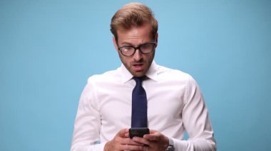 Genç yakışıklı iş adamı telefonunda gördüklerinden şok oluyor, yüzüne dokunuyor ve kafasını mavi arka planda sallıyor.