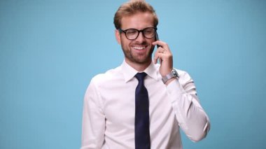 Yakışıklı iş adamı geniş gülümsüyor, telefonda konuşuyor ve mavi arka planda başparmak işareti yapıyor.