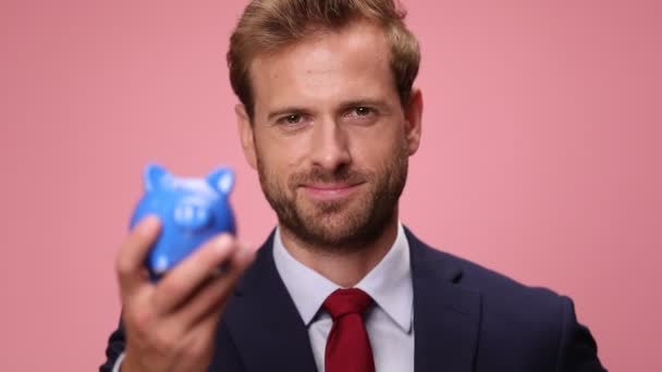 ネイビーブルーのエレガントな若いビジネスマンピンクの背景に家のモデルと貯金箱を示す — ストック動画