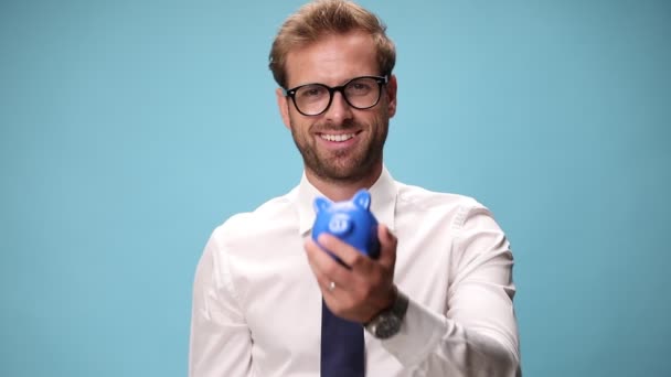 戴眼镜的商人面带微笑 拿着储蓄罐 把钱放在储蓄罐里 指着储蓄罐 在蓝色的背景上签了个名 — 图库视频影像