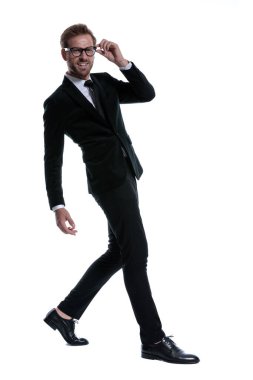 Siyah takım elbiseli gülümseyen genç adam, gözlük takıyor, kollarını moda pozunda tutuyor ve beyaz arka planda tek başına yürüyor.