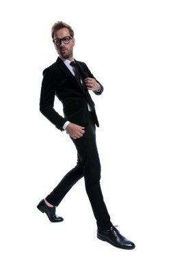 Dramatik havalı iş adamı kendine güvenerek siyah takımı ayarlıyor ve beyaz arka planda izole edilmiş bir şekilde, tüm vücudunu bir kenara atıyor.