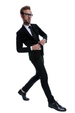 Siyah takım elbiseli dramatik genç adam ceketini ilikliyor ve beyaz arka planda şık bir şekilde yürüyor.