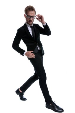 Siyah takım elbiseli ciddi bir adam, el ele tutuşuyor, gözlükleri tamir ediyor, aşağı bakıyor ve beyaz arka planda şık bir şekilde yürüyor.